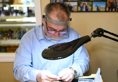 Leo Poisson closely examining a piece of jewelry with a jeweler's loupe on his head and a lamp beside him.