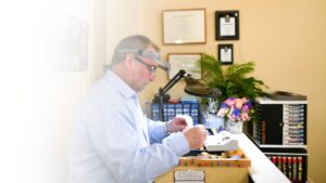 Leo Poisson, dressed in a light navy blue shirt, is seated in an office with organized files, various objects, wall decor, a plant behind him, and a bouquet of flowers in the corner of his desk. Wearing a watch and a jeweler's loupe on his head, he is intently focused on repairing a piece of jewelry under the intense white light of a black lamp.
