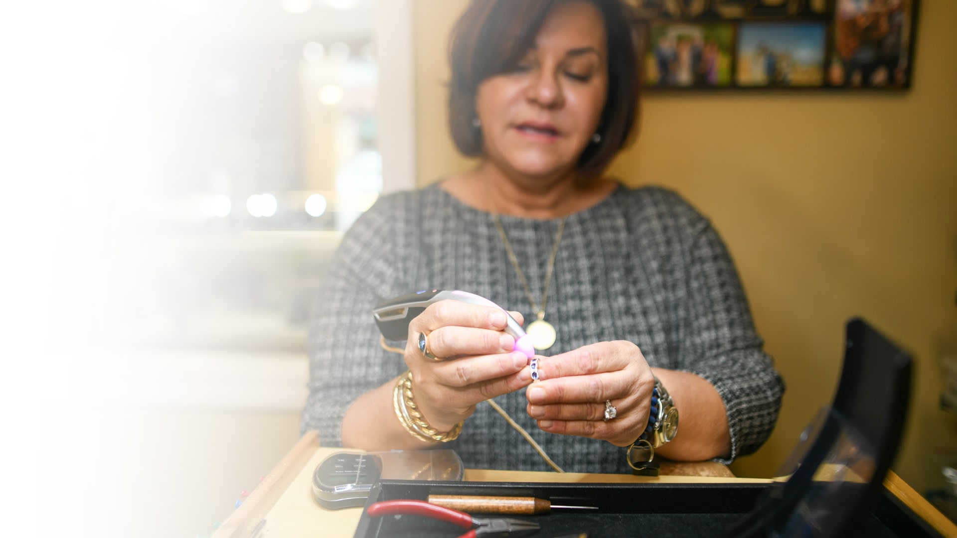 Lisa Poisson, dressed in a tweed suit, wearing a chain with a medallion, three bracelets, and a blue stone ring on her right hand, a watch, a blue bracelet, and a diamond ring on her left hand, examining another ring with multiple blue stones.