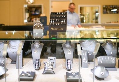 Earrings, necklaces, rings, and bracelets are delicately displayed on shelves with busts and cushions holding them, all in dark colors, with Leo Poisson in the background working on other shelves.