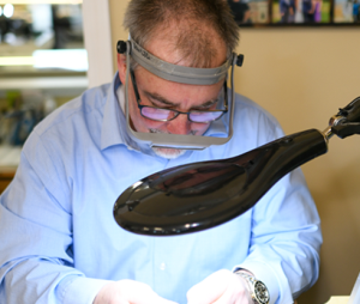 Leo Poisson closely examining a piece of jewelry with a jeweler's loupe on his head and a lamp beside him.
